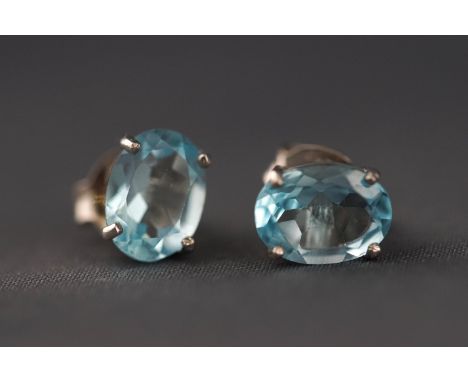 A white metal pair of single stone stud earrings each set with an oval faceted cut blue topaz. Post and butterfly fitting. St