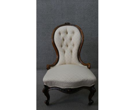 A Victorian walnut spoon back nursing chair, with a buttoned back, upholstered in patterned ivory fabric, on carved scrolling