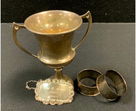 A George V silver twin handled trophy cup, Sheffield 1931;  a pair of silver napkin rings;  a decanter label 133.1g gross (4)