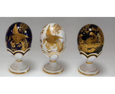 A set of three Royal Crown Derby Fabergé eggs, with stands, Gold Aves pattern, Black Aves pattern and Gold Aves pattern on Co