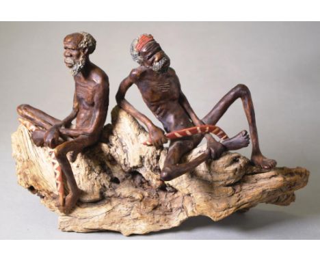 Australian ceramic figure of two indigenous men two men seated, each with a boomerang, on natural wood base. (As inspected: n
