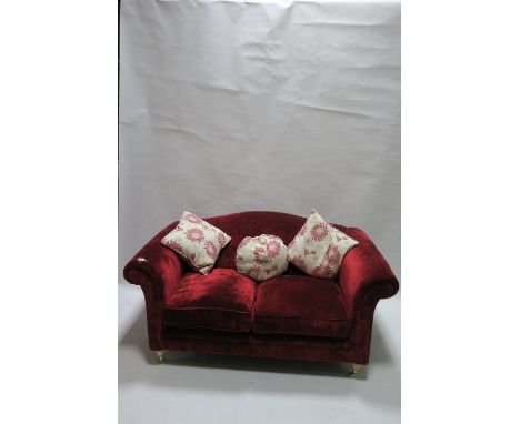 A RED VELVET UPHOLSTERED TWO SEATER SOFA, the hump back and shaped arms fitted with two loose seat cushions raised on circula