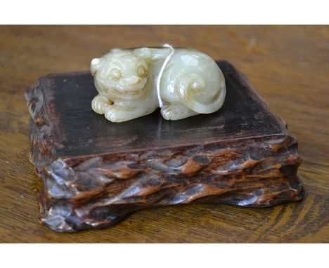 A Chinese pale celadon and russet jade carving, of a curled temple lion, 6cm wide, hardwood stand