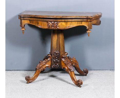 A Victorian rosewood card table of serpentine outline with red baize lined folding top, frieze carved with leaf scroll orname