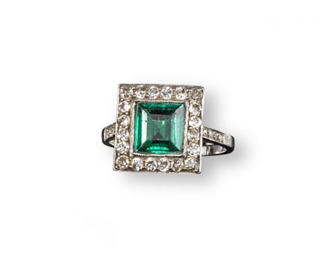 An emerald and diamond cluster ring, the square~shaped emerald is set within a surround of small circular~cut diamonds in pla