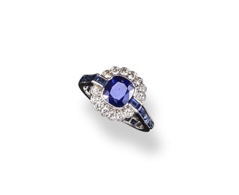 A sapphire and diamond cluster ring, the cushion~shaped sapphire is set within a surround of circular cut diamonds and flanke
