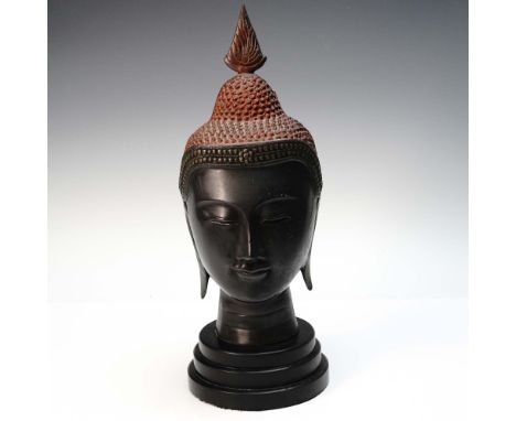 A cast bronze sculpture depicting the head of the Buddha, mounted on a black circular three-tiered pedestal base. The head is