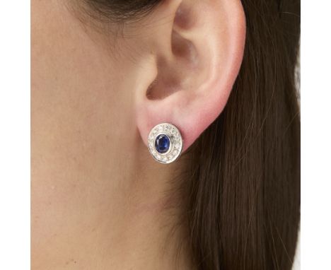 Pair of 14k white gold earrings, each bezel set with central oval blue sapphire and 13 full cut diamond halo. The two sapphir