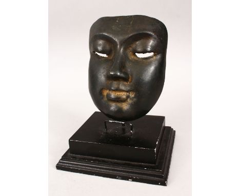 AN EASTERN BRONZE MASK with dark patination on a wooden base, 22cm high on stand, the mask 16cm high x 14cm wide.