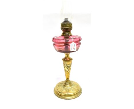 AN EDWARDIAN OIL LAMP  with cranberry glass reservoir and brass base with Art Nouveau relief decoration