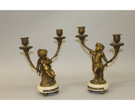 A GOOD PAIR OF 19TH CENTURY LOUIS XVI PATTERN CUPID BRONZE TWO LIGHT CANDLESTICKS on white circular marble bases 12ins high.
