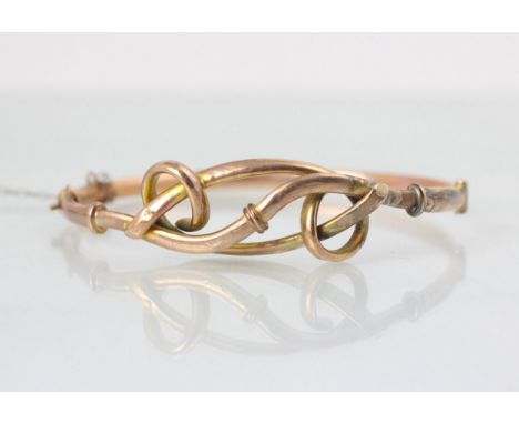 A yellow metal bangle, the hollow bangle with openwork bow style front, stamped ‘9ct’ suspending a yellow metal heart shaped 