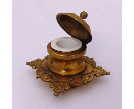 A brass inkwell shaped square, chased with scrolls and leaves the cylindrical well with porcelain liner, circa 1900, 9 x 9.5 