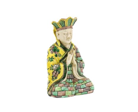 ~ A Chinese Porcelain Figure of an Aesthetic, Kangxi, seated crossed legged, his hands in prayer, wearing a lappet-moulded ca