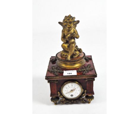 An early 20th century mantel clock with an enamel dial in a wooden faux marble case, crowned by a metal cupid reading a scrol