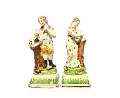 A pair of Staffordshire figures of children, c.1800, each standing beside a tall stump, the boy with a dog, the girl with a l