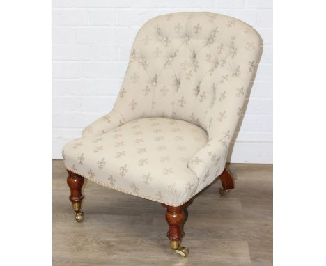 An antique button back low chair with fleur-de-lys upholstery, turned legs and brass castors, 52cm wide x 70cm tall