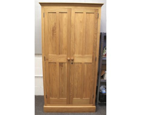 A contemporary light oak wardrobe. Two panelled doors with turned handles, opening to reveal a clothes rack, on pedestal base