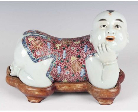 A Chinese famille rose porcelain pillow, 20th century, modelled as a boy resting on his knees and elbows with head turned to 