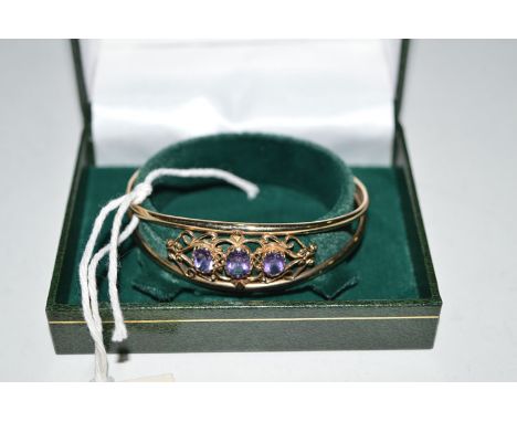 A 9ct. yellow gold and amethyst set bangle, the three graduated oval facet cut amethysts within scrolling mount.