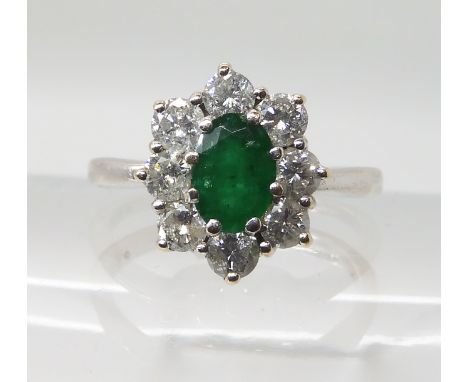 A 9CT WHITE GOLD EMERALD AND DIAMOND CLUSTER RING set with an oval emerald of approx 7mm x 5mm x 3.6mm, the estimated approx 