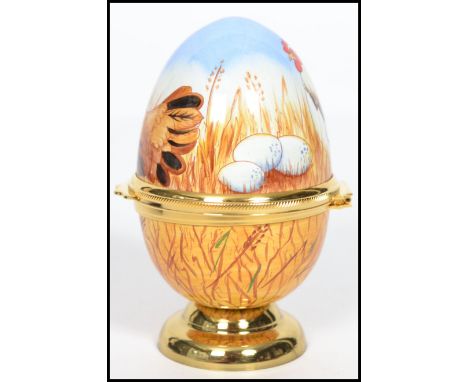 A Moorcroft painted enamel on copper trinket box in the form of an egg, decorated with chickens, eggs and hay, the gilt metal