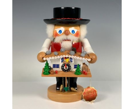 A handmade wooden figure of a man holding a cuckoo clock, wearing a red vest and black hat adorned with a silky red rope. Ste