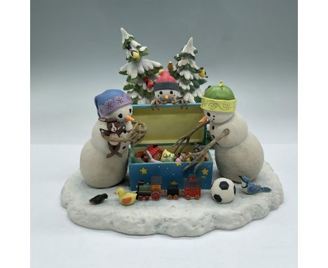 This illuminated sculpture depicts three snow children digging through an open toy chest. Two snowy trees stand in the backgr