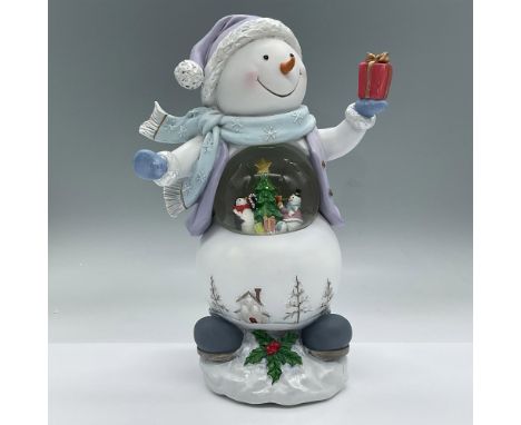 A cheerful snowman dressed in a lavender hat and sweater holding up a present has a snow globe in his midsection. Inside the 