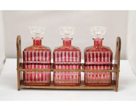 A WOODEN HOLDER CONTAINING THREE AUSTRIAN STYLE 1930'S RUBY RED FLASHED ART DECO GLASS DECANTER LIQUOR BOTTLES 