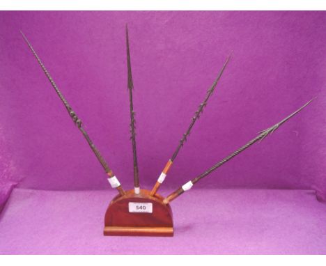 Four early 20th century metal fishing arrow heads having different barbed tips, in mahogany stand