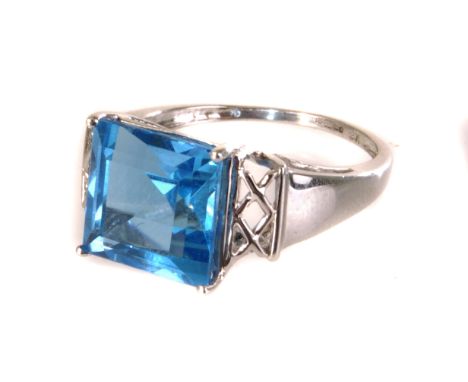 A 9K white gold and topaz dress ring, the square cut blue stone set in four claw mount, with pierced crosshatched design to s