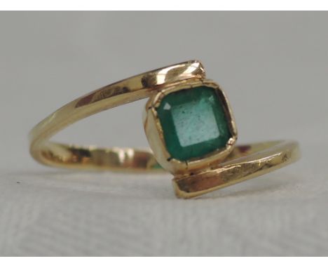 A lady's dress ring having a single square emerald in a collared mount to crossover shoulders on a yellow metal loop, no mark