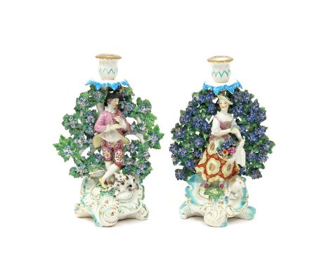 A pair of Derby candlestick figures of a shepherd and his companion  c.1765-68, both standing before flowering bocage, he pla