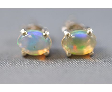 A white metal pair of single stone stud earrings, each set with an oval cabochon cut Ethiopian opal. Post and scroll fittings