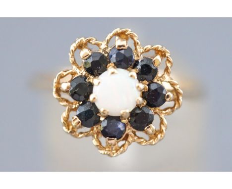 A yellow metal floral design cluster ring set with a central round cabochon opal and surrounded by dark blue sapphires. Size 