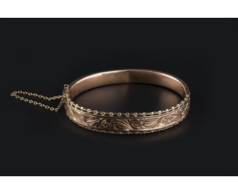 A 9CT GOLD BANGLE, of hinged oval form, engraved with foliate scroll decoration between scalloped borders, inner diameter 5.8