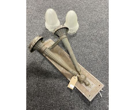 An antique patinated brass twin branch wall light in the form of torches, with opaque glass shades, mounted on brass plate 
