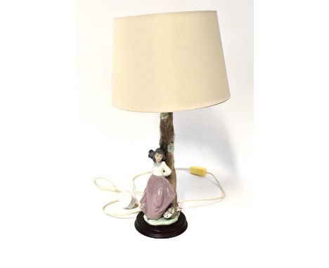 LLADRÓ; a figural lamp base with a young lady with a basket of flowers by her feet and leaning on a tree, raised on circular 
