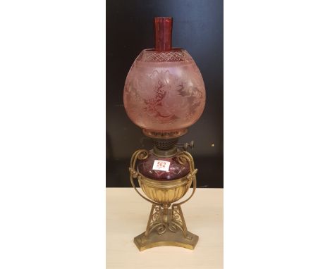 An Edwardian brass and cranberry glass oil lamp: standing on tri-form base, shade and chimney present, overall height 56cm. 