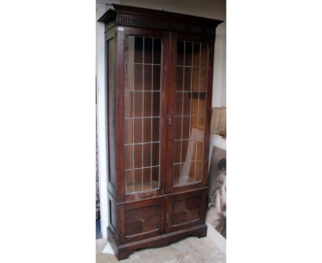 AN EDWARDIAN OAK DISPLAY CABINET with lead glazed doors, further cupboard doors beneath and standing on bracket feet (shelves