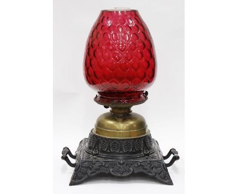 AN EARLY VICTORIAN CAST IRON OIL LAMP with cranberry glass shade which has a honeycomb decoration, 50cm high 