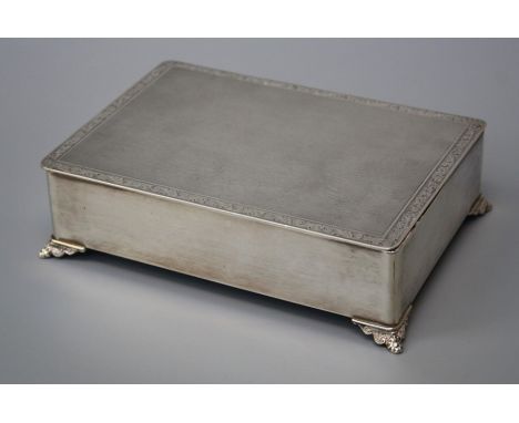 A silver cigarette box, rounded rectangular engine turned with foliate engraved border, hinged cover and foliate bracket feet