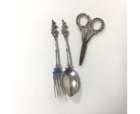 A white metal fork and spoon, both with rabbit finials and a pair of silver handled scissors, handles in a rococo style (11cm
