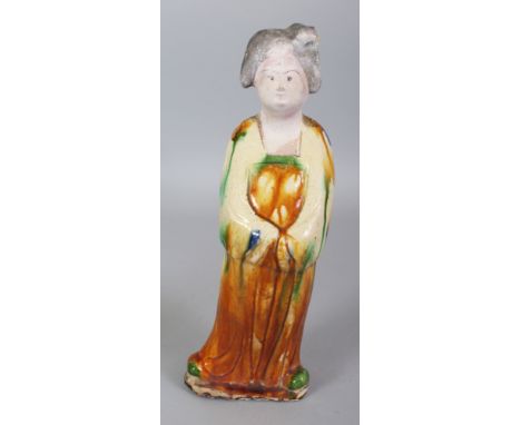 A CHINESE TANG STYLE GLAZED CERAMIC FIGURE OF A STANDING LADY, her robes glazed in ochre and green, her unglazed face and hai