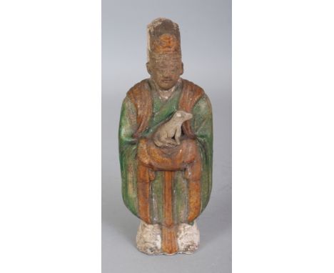 A CHINESE TANG STYLE GLAZED CERAMIC FIGURE OF A STANDING ATTENDANT, bearing a dog in his arms, the figure applied with green 