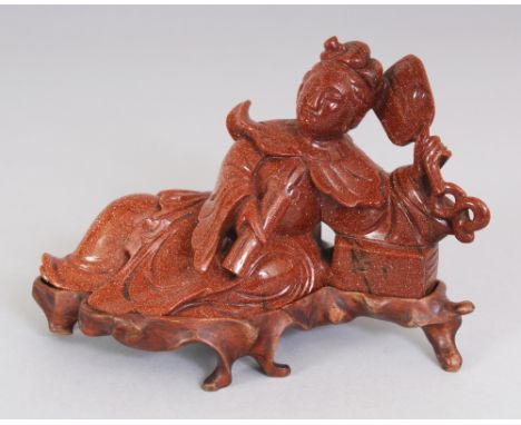 A 20TH CHINESE GOLDSTONE MODEL OF A RECLINING LADY, together with a fitted wood stand, the lady holding a fan and a closed sc